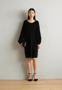 Black long-sleeve sweater dress with a round neckline. Smooth texture, knee-length, paired with black high-heeled shoes and a small handbag.