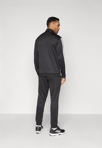 Čierny športový tréningový súprav s bielymi pruhmi na bokoch, opatrený zipsovým golierom a elastickými manžetami. Nosený s čiernymi športovými topánkami.