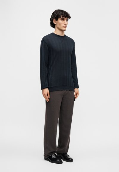 Μπλε ναυτικό πουλόβερ με υφή και σχέδιο ψαροκόκαλο, σε συνδυασμό με γκρι παντελόνι. Το μοντέλο στέκεται δίπλα με τα χέρια στις πλευρές, φορώντας μαύρα παπούτσια.