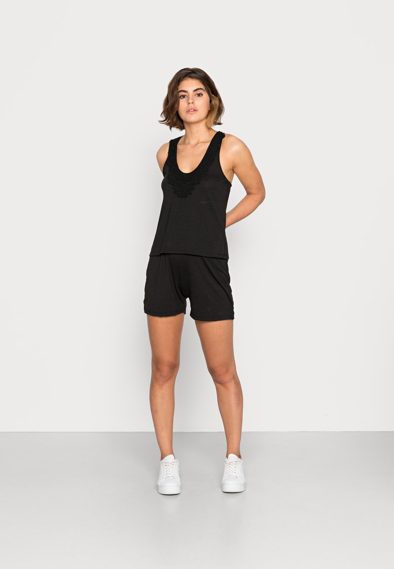 Top preto sem mangas com decote bordado, combinado com calções pretos e usado com ténis brancos. Design simples, textura suave e estilo casual.