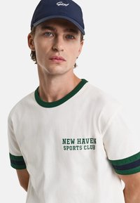 White cotton T-shirt with green and navy striped accents, featuring "NEW HAVEN SPORTS CLUB" printed in green on the front.