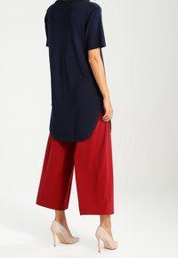 Camicia tunica blu navy a maniche corte con orlo curvo abbinata a pantaloni larghi rossi e tacchi beige, con attenzione al contrasto di colori e alla vestibilità.