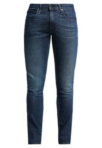 Tmavě modré slim-fit džíny s pěti kapsami, zapínání na knoflík a s mírným vyšisováním na předních stehnech a kolenou, vystavené na bílém pozadí.