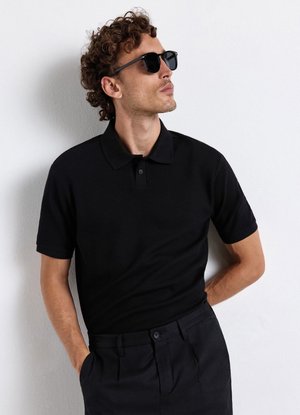 Mann mit lockigem Haar, der schwarze Sonnenbrille, schwarzes Poloshirt mit kurzen Ärmeln und schwarze Hose trägt, steht an einer weißen Wand.
