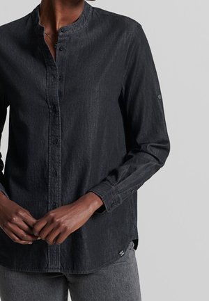 Personne portant une chemise noire à manches longues avec col mandarin, les mains jointes près de la taille, debout devant un fond clair uni.