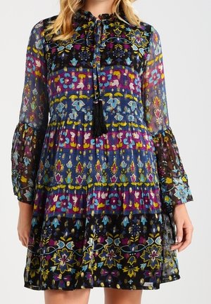 Sukienka z długim rękawem z przezroczystej tkaniny, w wielokolorowe kwiatowe i geometryczne wzory, z falbankowym kołnierzem oraz czarnymi frędzlami przy dekolcie.