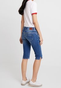 Jeans in denim blu slim fit con vita media e taglio all'altezza del ginocchio, caratterizzati da una piccola fessura all'orlo e una patch in pelle marrone con il logo sul retro.