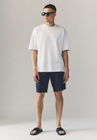 Valge üle pikkade varrukatega T-särk, mereväe värvi lühikesed püksid, mustad sandaalid ja tumedad päikeseprillid. Sile kangas, lõdvestunud lõige ja minimalistlik disain.