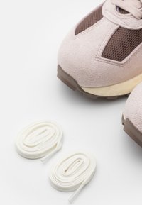 Jasnoróżowy zamszowy sneaker z brązowymi panelami z siateczki, obok na białym tle znajdują się białe, okrągłe sznurowadła.