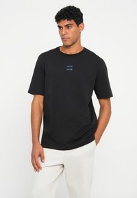 Vīrietis ar melnu īsu piedurkņu t-kreklu, uz krūšu daļas ar maziem ziliem uzrakstiem un gaišiem biksēm, stāv uz vienkārša baltā fona.