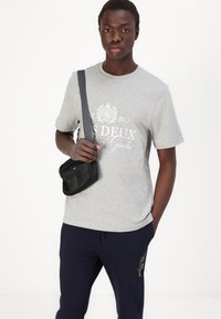 Grijze t-shirt met korte mouwen en witte opdruk, gecombineerd met marineblauwe trainingsbroek en een zwarte schoudertas met een riem.