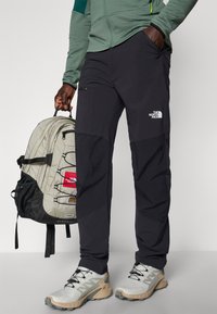 Svarte utendørsbukser med glidelåslommer, kombinert med en grå North Face ryggsekk med flere rom og en nettinglomme. Grå sko.