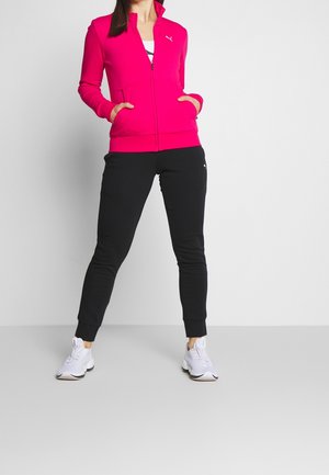 Woman wearing a bright pink zip-up jacket, black jogger pants, and white sneakers, standing with hands in pockets against a plain background.
