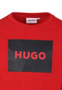 Rode trui met een zwart rechthoekig vlak waarop het woord "HUGO" in rood is afgebeeld. Ribbelhals en een zachte textuur, gemaakt van katoen.