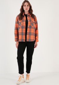 Narancssárga kockás ingdzseki fehér gombokkal, két mellzsebbel, fekete nadrággal és narancssárga díszítéssel ellátott fehér tornacipővel párosítva.