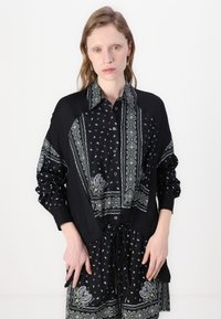 Svart skjorte med intrikate paisley- og geometriske mønstre i hvitt og grønt, lange ermer og beltedetalj med snøring.