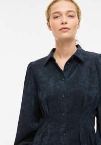 Tamnoplava haljina od kordurove tkanine s ovratnikom, prednjim dijelom na kopčanje i naboranim strukom. Ima duge rukave i suptilnu teksturu.