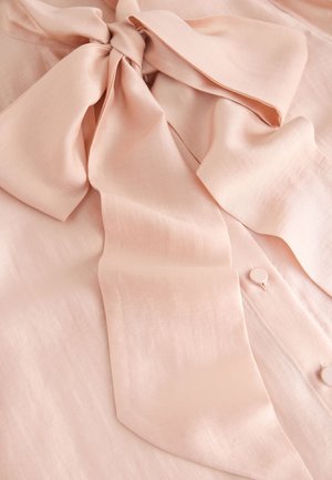 Jasnoróżowa bluzka z gładkiej tkaniny, z dużą kokardą przy dekolcie i guzikami pokrytymi materiałem.