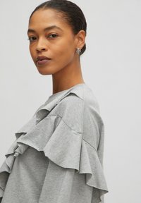 Grauer Pullover mit geschichteten Volants an den Ärmeln, aus weichem Stoff. Das Model trägt silberne Creolen und hat eine glatte Frisur nach hinten.