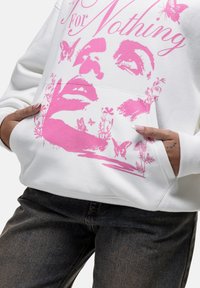 Witte hoodie met een roze grafische print van een gezicht, vlinders en bloemen. Inclusief een buidelzak en geribde manchetten, gecombineerd met donkere jeans.