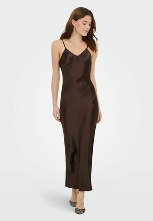 Vrouw draagt een donkerbruine satijnen slipdress met dunne bandjes en hoge hakken met luipaardprint, staand tegen een effen achtergrond.