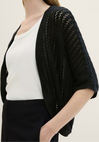 Zwarte opengebreide cardigan met een gestructureerd patroon, voorzien van korte mouwen en een losse pasvorm, gedragen over een witte nauwsluitende top.
