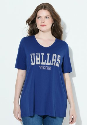 Mujer con cabello largo castaño que lleva pendientes de aro plateados y una camiseta azul de cuello en V con el texto "DALLAS TEXAS", combinada con jeans azules.