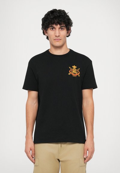 T-shirt en coton noir à manches courtes, avec un emblème brodé en or sur la poitrine gauche. Design uni, col rond, coupe décontractée.