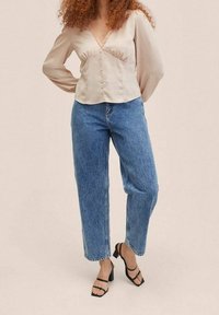 Femme portant une blouse beige à manches longues avec des boutons, un jean taille haute bleu et des talons blocs noirs à lanières, se tenant devant un fond uni.