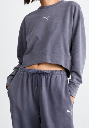 Osoba ubrana w wyblakłą, ciemnoszarą skróconą bluzę i pasujące spodnie dresowe z logo Puma, ręce w kieszeniach, na jednolitym jasnym tle.