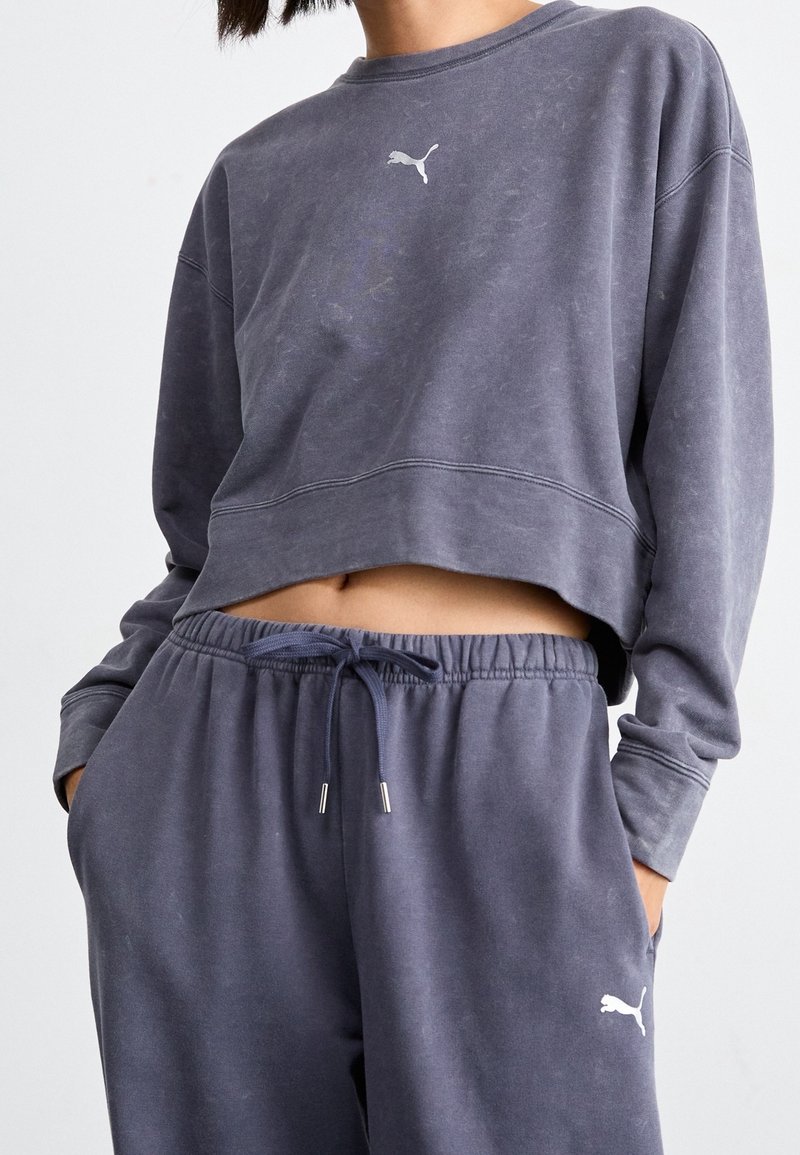 Personne portant un sweat-shirt court gris foncé délavé et un jogging assorti avec logos Puma, mains dans les poches, sur un fond clair uni.