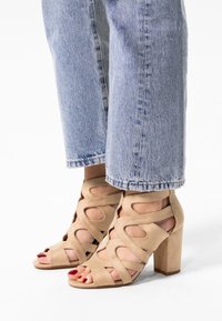 Beige hoge sandalen van suede, met uitsnijdingen en een vierkante hak. Draagt het met opgerolde lichtblauwe jeans.