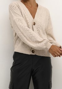 Béžový pletený kardigan s výstřihem do V, texturovaným chevronovým vzorem, velkými hnědými knoflíky a žebrovanými manžetami. Nositý přes černé kožené kalhoty.