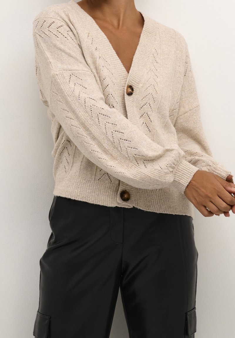 Béžový pletený kardigan s výstřihem do V, texturovaným chevronovým vzorem, velkými hnědými knoflíky a žebrovanými manžetami. Nositý přes černé kožené kalhoty.