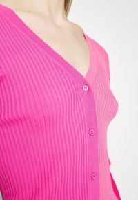 Fitted, ribbed cardigan in pink, featuring a V-neckline and five pink buttons down the front. Soft texture and seamless design.