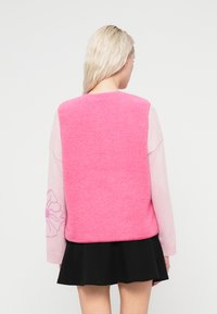 Ροζ αμυγδαλωτό sherpa γιλέκο με μακριά ροζ μανίκια διακοσμημένα με περίγραμμα λουλουδιού. Ολοκληρώνεται με ένα τελείωμα που βρίσκεται πάνω από μια μαύρη φούστα.