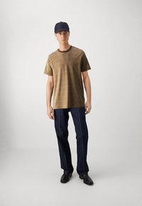 Ruskea kuvioitu t-paita pyöreällä pääntiellä, yhdistetty tummansinisillä suorilla farkuilla ja mustilla saappailla. Mallilla on päässään tummansininen baseball-lippis.