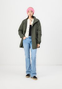 Parka vert olive avec un cordon de serrage à la taille, un pull beige clair à boutons, un haut noir, un jean évasé bleu et un bonnet en tricot rose.