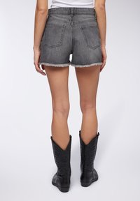 Bermuda in denim nero sbiadito con orli sfrangiati e due tasche posteriori, abbinati a stivali cowboy neri fino al ginocchio con cuciture decorative.