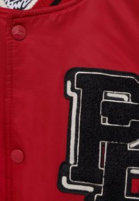 Veste rouge avec un écusson en forme de lettre "P" noire texturée, bordé de blanc. Comprend un bouton pression rouge et un col côtelé.