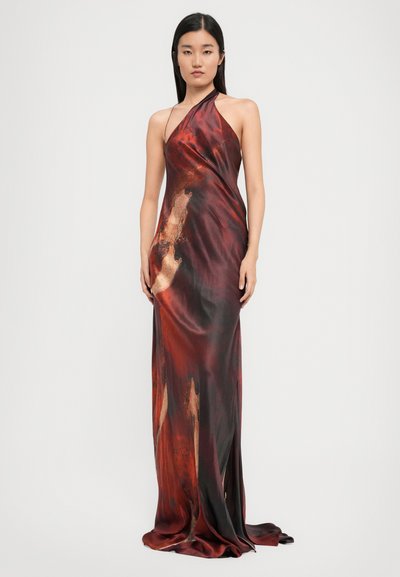 Woman standing wearing a long, sleeveless halter dress with a red, brown, and black abstract pattern against a plain white background.
