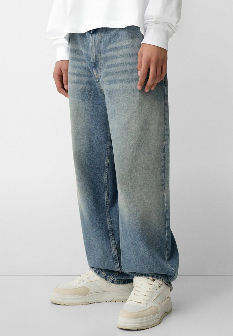 Helle blaue weit geschnittene Jeans mit ausgewaschenem Look, hoher Taille und kontrastierendem Nähen, kombiniert mit weißen Sneakers.