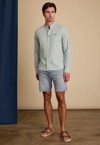 Lichtgroen lang mouw shirt met een bandkraag en knoopdetails, gecombineerd met blauwe denim shorts en bruin sandalen op een effen achtergrond.