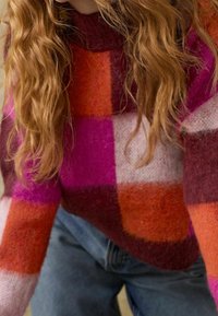 Bunt gestrickter Pullover mit großen Farbkombinationen in Pink, Orange und Bordeaux. Hoher Kragen und weiche Textur. Kombiniert mit Jeans.