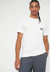 Tricou alb din bumbac, cu mâneci scurte, guler rotund și un buzunar pe piept cu o etichetă din material albastru și alb. Poartă pantaloni scurți gri.