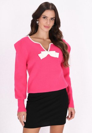 Roze gebreide trui met een V-hals, witte rand en een grote witte strik op de borst. Zwarte aangesloten rok. Lange mouwen met ribbelmanchetten.