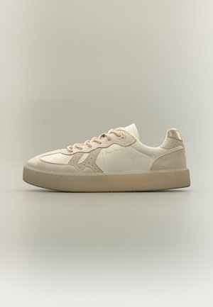 Baskets basses beige et crème avec détails en daim, design perforé sur le côté, lacets et semelle plate en caoutchouc sur fond neutre.