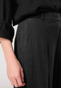 Schwarze Leinenhosen mit taillierter Passform, ausgestattet mit Seitentaschen und einer glatten Textur. Neben einem teilweise sichtbaren dunklen Hemd gezeigt.