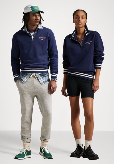 Námořnický sweatshirt na zip s bílými pruhy, v kombinaci se šedými tepláky a zelenými teniskami. Černé šortky a sportovní černé boty na modelu.