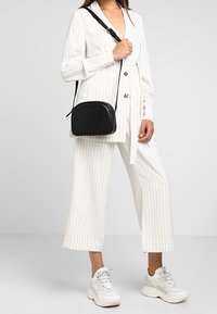 Borsa a tracolla nera con forma curva, in pelle liscia e con tracolla regolabile, indossata su un outfit bianco a righe con pantaloni larghi.